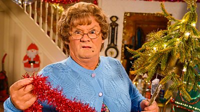 Brendan O'Carrol's Mrs Brown stares down the camera making a face while holding tinsel, standing beside a Christmas tree.