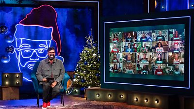 Romesh Ranganathan sits in front of a large neon light depicting him as Santa while his Ranganation appear on screen beside him