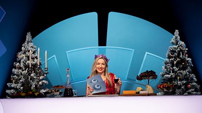 Only Connect host Victoria Coren poses in a Christmas hat, holding a glass with a cheeseboard on her desk.