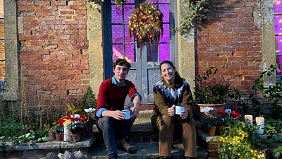 Arthur Parkinson and Rachel De Thame sit on a step holding cups. A festive wreath hangs on the door behind them.