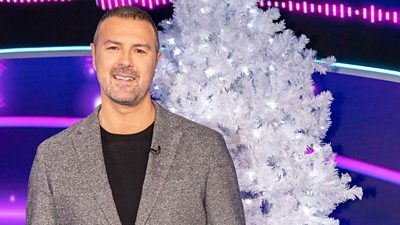 Paddy McGuinness stands in front of a bright white and silver Christmas tree.