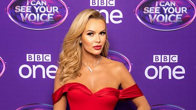 Amanda Holden wears a red dress and stands in front of a purple board.