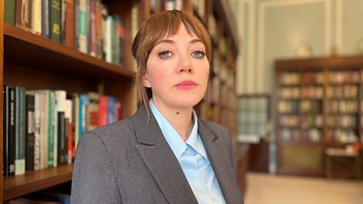 Philomena Cunk stands in a library, surrounded by shelves of books