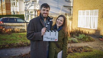 Tom Burke as Cormoran Strike and Holliday Grainger as Robin Ellacott, holding a clapperboard