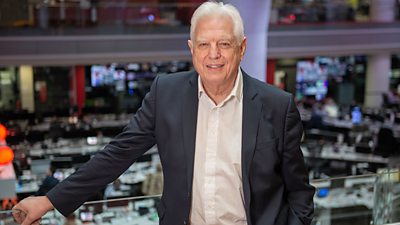 John Simpson stands against the backdrop of the BBC newsroom wearing a dark suit jacket over a light shirt.