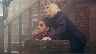 Yasmin Khan (Mandip Gill) and The Doctor (Jodie Whittaker) hide behind a barrel on a ship.