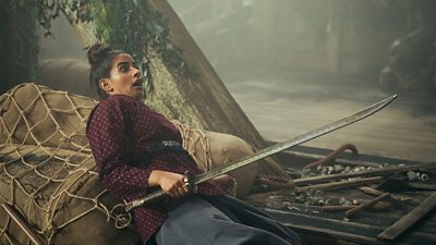 Yaz (Mandip Gill) lies on top of a pile of sacks on board a ship with a sword in her hand. She looks frightened.