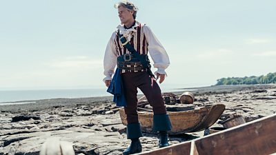 Dan (John Bishop) stands on the shoreline in the puff sleeved shirt and flared trousers of a pirate