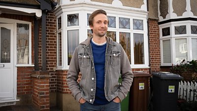 Tom Basden stands outside a red brick house with white windows and doors and wheelie bins in the front garden