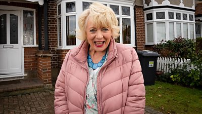 Alison Steadman stands outside a red brick house with white windows and doors and wheelie bins in the front garden