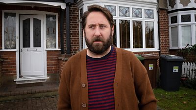 Jim Howick stands outside a red brick house with white windows and doors and wheelie bins in the front garden