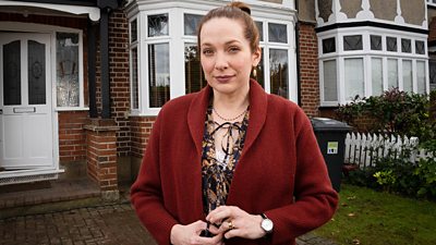 Katherine Parkinson stands outside a red brick house with white windows and doors and wheelie bins in the front garden