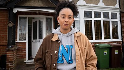 Mica Ricketts stands outside a red brick house with white windows and doors and wheelie bins in the front garden