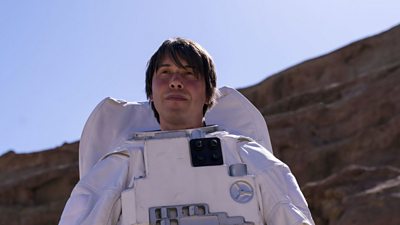 Professor Brian Cox wears an astronaut's white suit, without the helmet.