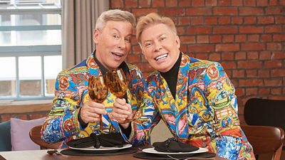 Royston and Nick wear brightly coloured matching patterned suits and clink their glasses in a cheers gesture at the dinner table