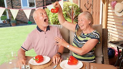 Keith and Linda Bailey sit in the back garden at a table. She holds some tomatoes above his open mouth. They each have a red pepper on their dinner plates.