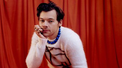 Harry styles wears a white fluffy jumper with a pattern of a house on it. He wears a blue beaded necklace and rests his face in his hand. He wears multiple rings and sits in front of a red curtain.