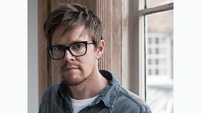 Kris Marshall wears dark framed glasses, a white t-shirt and denim shirt. He stands by a window.