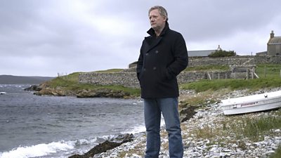 DI Jimmy Perez (Douglas Henshall) stands on the rocky coastline looking out to sea. He wears a black coat and jeans.