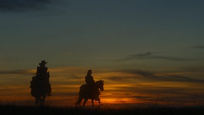 Two figures ride on horseback in silhouette against the backdrop of the setting sun