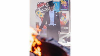 Neil Patrick Harris stands behind a burning item in a top hat and tails