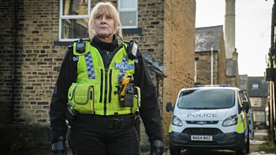 Sarah Lancashire as Catherine Cawood in Happy Valley. She stands in the road in a high vis police vest and uniform with a police van behind her.
