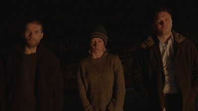 Man, woman and man standing looking at camera looking concerned.  Image is dark and ominous 