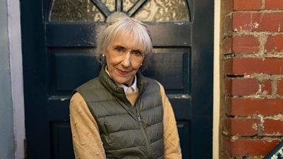 Anita Dobson's character stands outside a dark front door in a green bodywarmer and cream jumper.