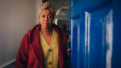 Michelle Greenidge stands at an open blue front door wearing a red coat and yellow cardigan