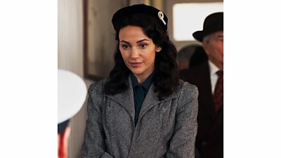 Michelle Keegan as Kate. She wears a caot and hat as she speaks to a man at a desk.