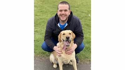 Robbie Crow smiles, crouching down with his arms around a Golden Labrador