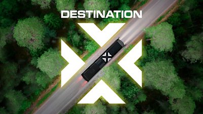 A black bus with an X on the trop drives along a road through a wooded area