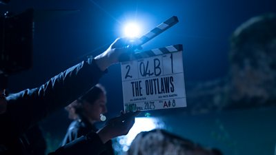 Hands hold a clapperboard on a night shoot for The Outlaws. A figure is visible in the background with light reflected on the ground.