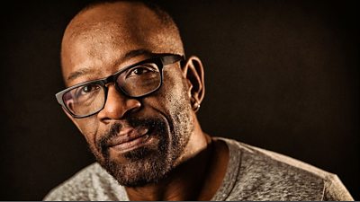 Lennie James wears a grey t-shirt and black-framed glasses, he is photographed against a plain black background