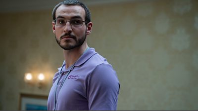 Éanna Hardwicke as Ben Field, he has a short dark beard and wears glasses, he is wearing a lilac coloured polo shirt