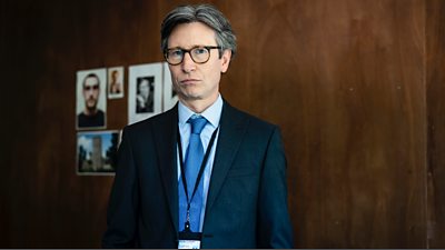 Jonathan Aris as DCI Mark Glover, a man with greying hair who wears glasses and a dark blue suit, he has a lanyard around his neck with a pass attached