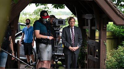 Timothy Spall as Peter Farquhar standing next to the camera crew in the entrance of a churchyard