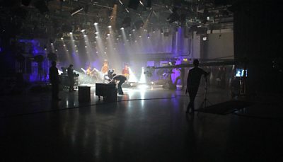 A large studio with a rock band playing. A variety of cameras and equipment are in the foreground. 