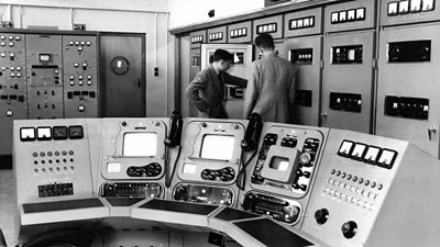 Two men examine a large rack of equipment. In front of them is a panel of monitors and dials. 