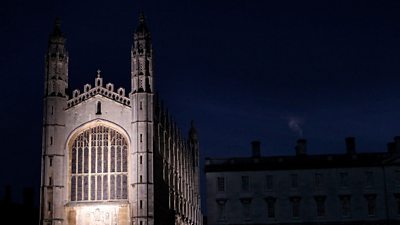 The exterior of King's College Cambridge at night. One side of the building is illuminated by a light on the ground. 