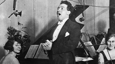 Mostyn Thomas in full white tie sings into a horn microphone. A woman at a piano smiles behind. 