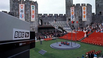 A tv television camera looks over the dais with three thrones at Caernarfon Castle which is festooned with heraldic banners