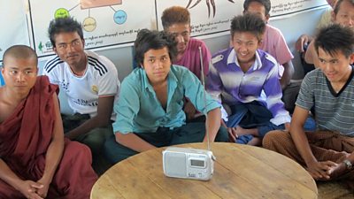 A group of people sit crossed legged gathered around a radio on a table.