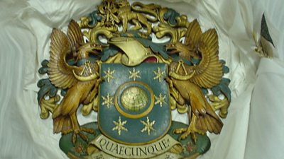 The ý coat of arms picked out in blue and gold. Two eagle supporters with horns hold a shield with a globe and seven stars. A lion holding a thunderbolt motif. The motto is Quaecunque. 