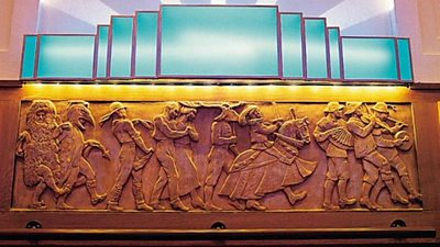 A frieze consisting of figures dancing in a procession in costumes and fancy dress. Above is an art deco light.