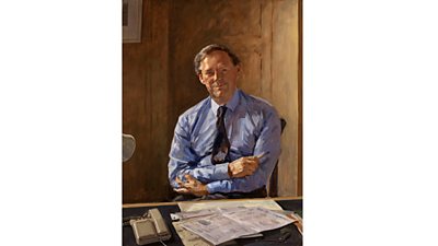 Portrait of Michael Checkland. He sits at his desk looking at the viewer with folded arms but a kind expression. Various papers and a 1990s pushbutton telephone are on his desk. 