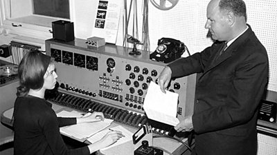Delia Derbyshire sits at a console with various levers, knobs and dials. Desmond Briscoe stands over her with a book in hand. 