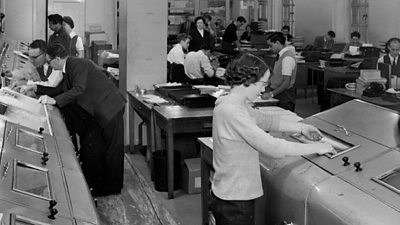 A busy scene of men and women operating various large pieces of equipment while journalists sit at desks in the background. 