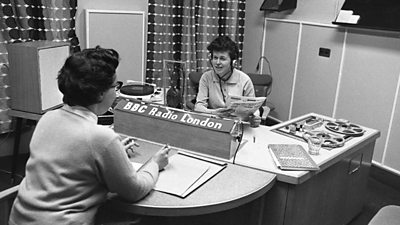 Two women sitting page-to-face discuss the newspapers in a studio. A reel-to-reel deck and a record deck can be seen. 