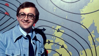 Ian McCaskill dressed quite informally in a blue denim jacket. He grins at the camera in his square glasses. 